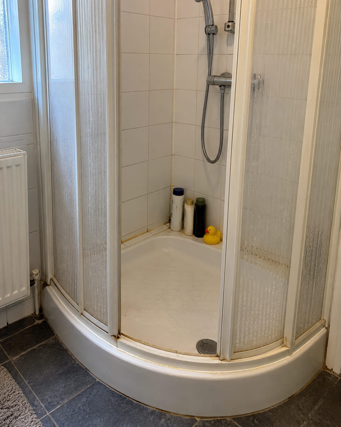 Voor de renovatie: een kwartronde kunststof douchecabine met douchebak en kleine witte wandtegels.
